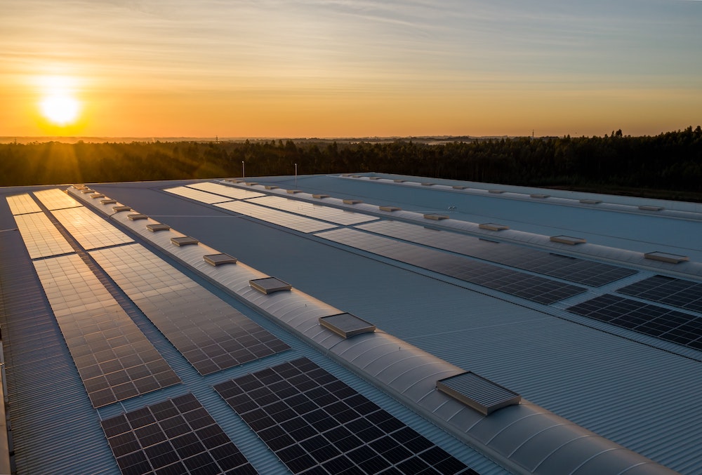 A beautiful sun rising over a white industrial roof that that rows of solar monitors and solar panels.